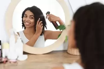 Young attractive black female using hair straightener products while sitting at a vanity and looking into mirror..