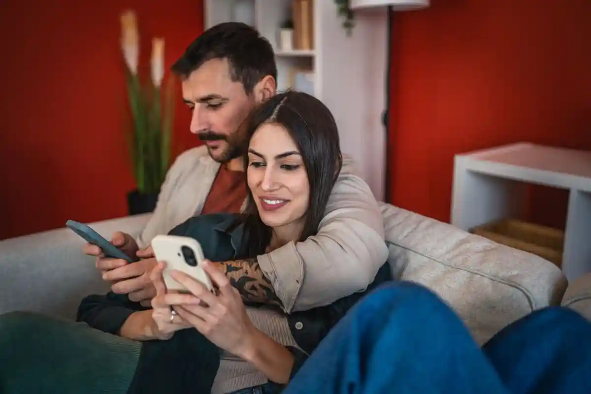 Family couple enjoy online leisure at home, use a mobile phone for entertainment and conversation. Together they explore the internet, hug a moment of connection and relaxation in home on sofa