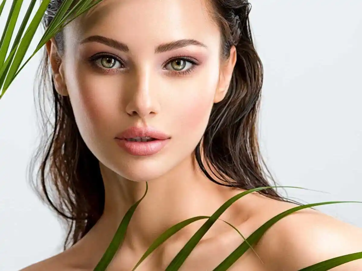 Young beautiful woman with healthy skin of face and palm leaves. Closeup fresh face of an attractive caucasian girl with green plants. Model with bright brown eye makeup. Skin care concept.

